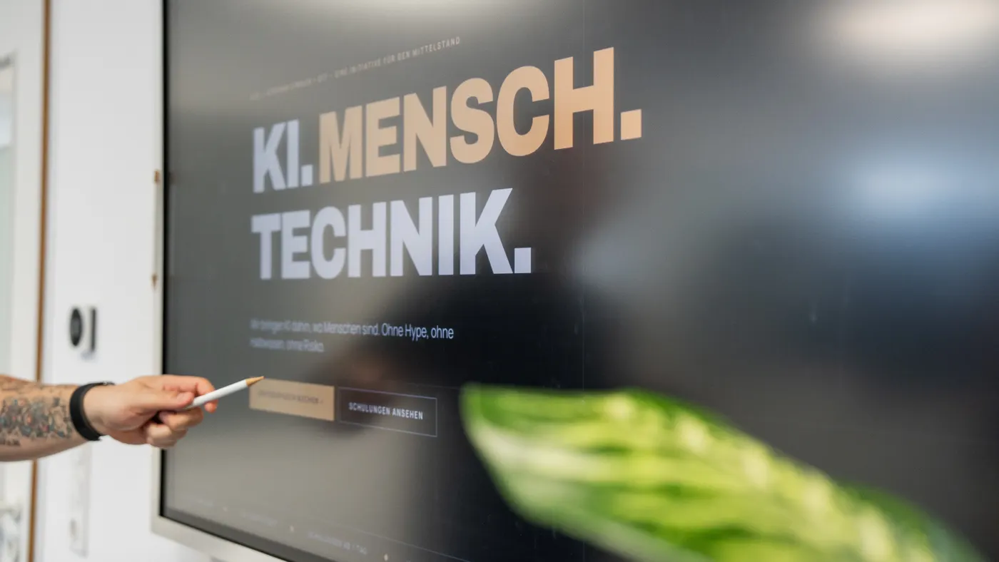 Bildschirm mit dem Schriftzug KI. Mensch. Technik., ein Stift zeigt darauf
